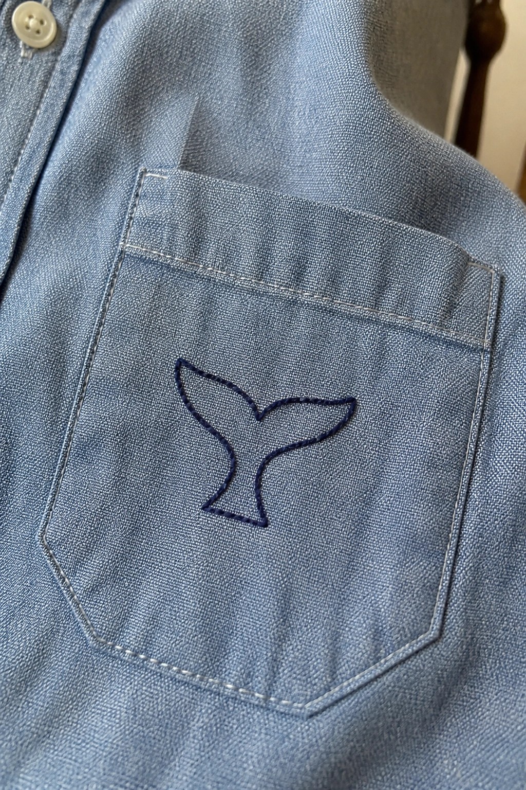 Light blue denim shirt pocket with navy embroidered whale tail design