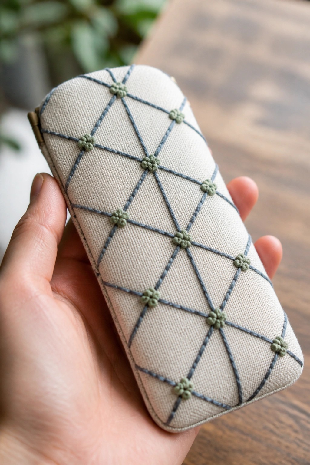 Hand holding beige pouch with gray diamond grid and green floral knots.