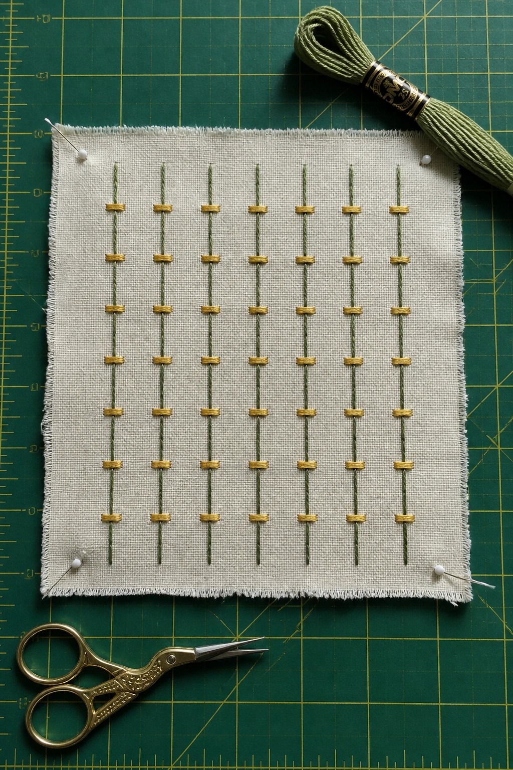 White fabric with green and gold grid embroidery pinned on mat beside scissors and thread