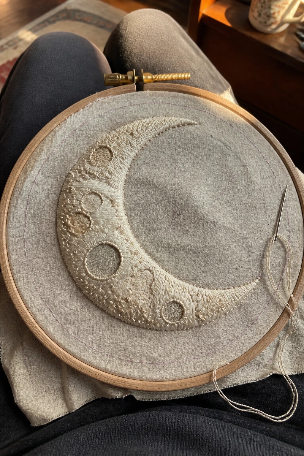 Wooden embroidery hoop with white crescent moon stitching and loose needle on lap.