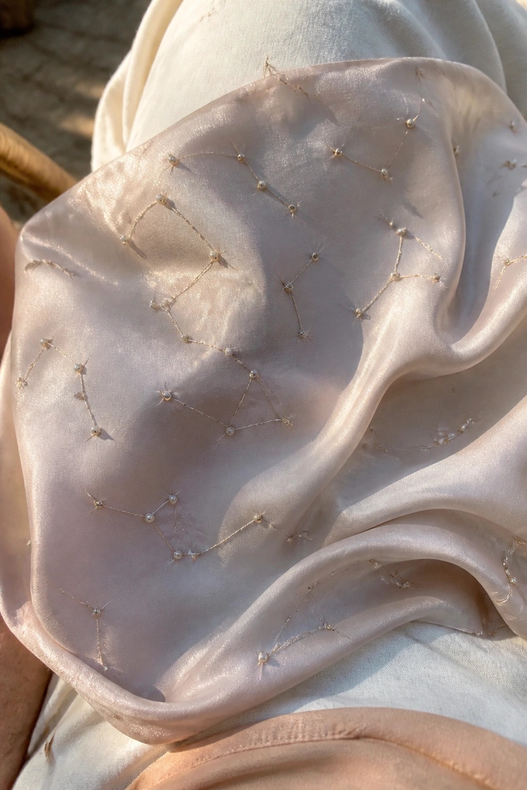 Light pink satin fabric with delicate gold thread and pearl embroidery draped softly in sunlight.