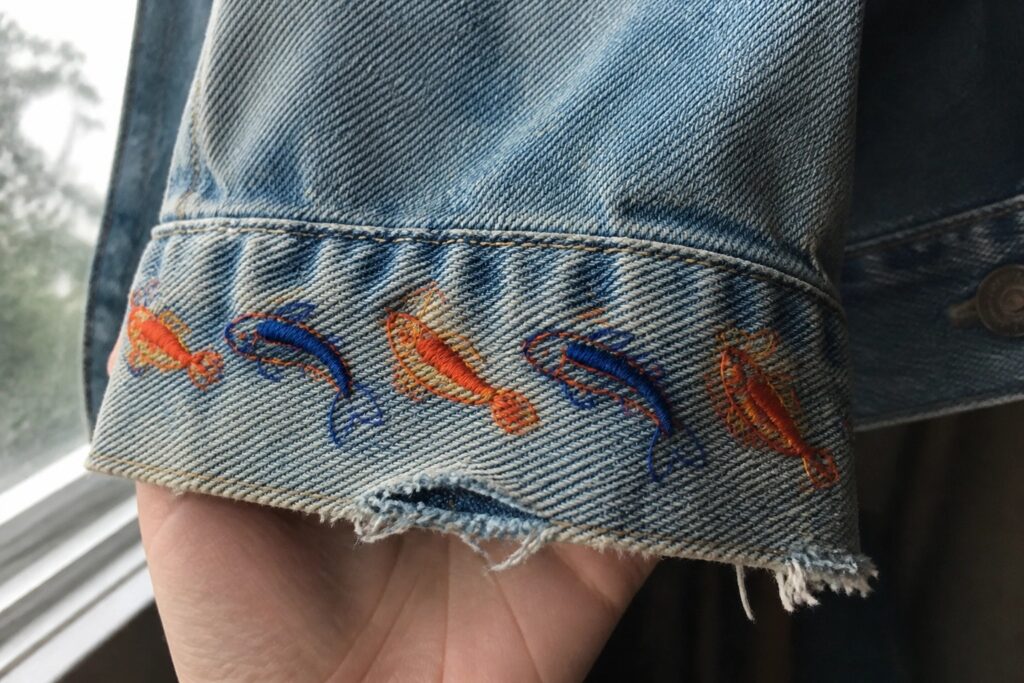 Hand holding light blue denim cuff embroidered with orange koi fish in waves.