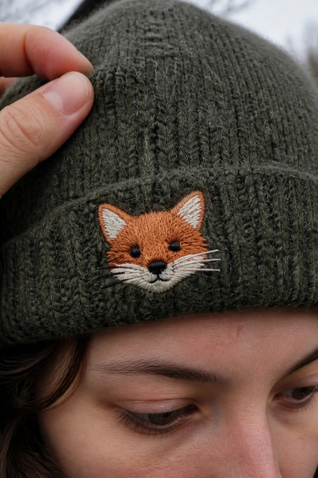 Green knitted beanie with orange embroidered fox on cuff, hand touching brim