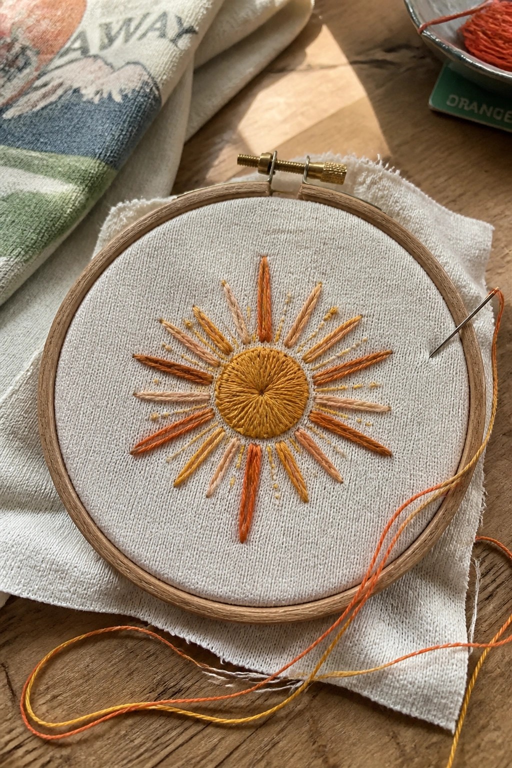 Orange sun embroidery in wooden hoop with needle and loose threads on white fabric.