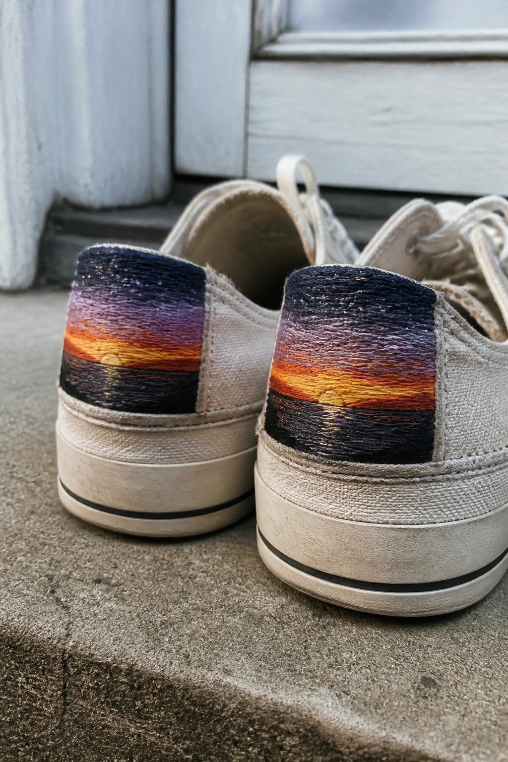 White canvas sneakers with colorful sunset embroidery on the heels, resting on concrete.