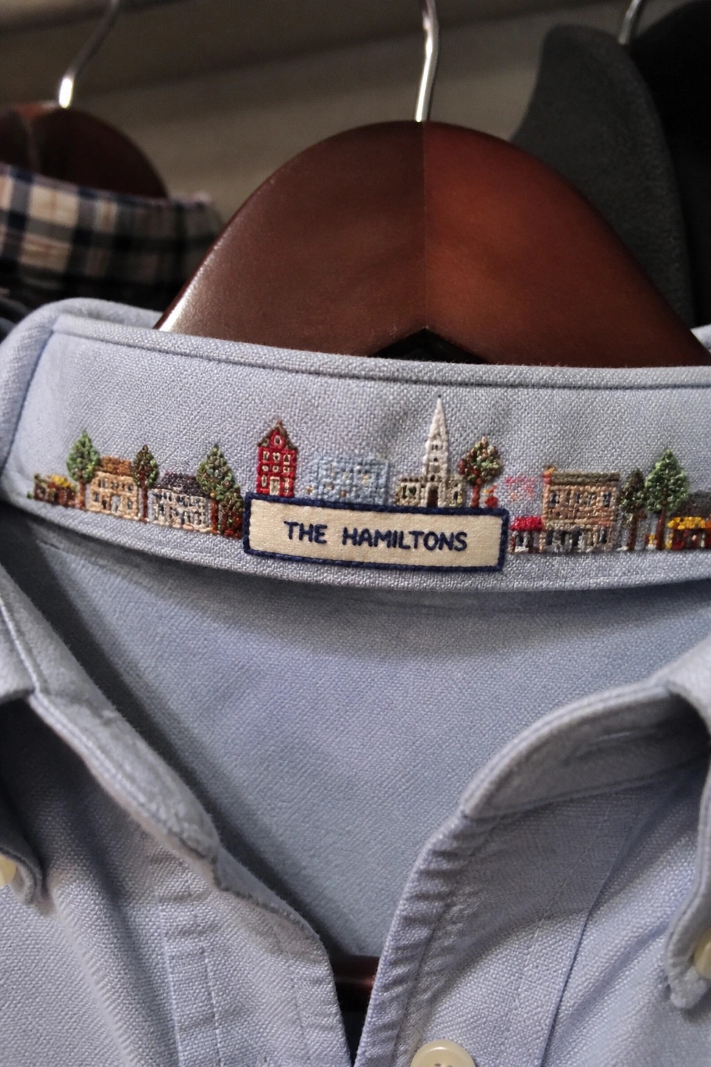 Light blue shirt collar with embroidered town buildings and trees above a name patch reading THE HAMILTONS.