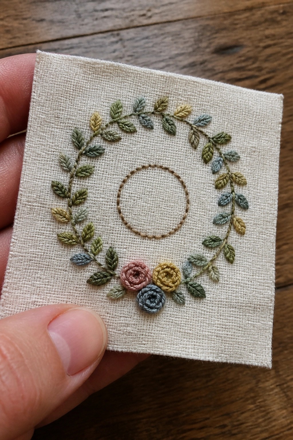 Hand holds linen with embroidered leaf wreath, central circle, and three small roses.