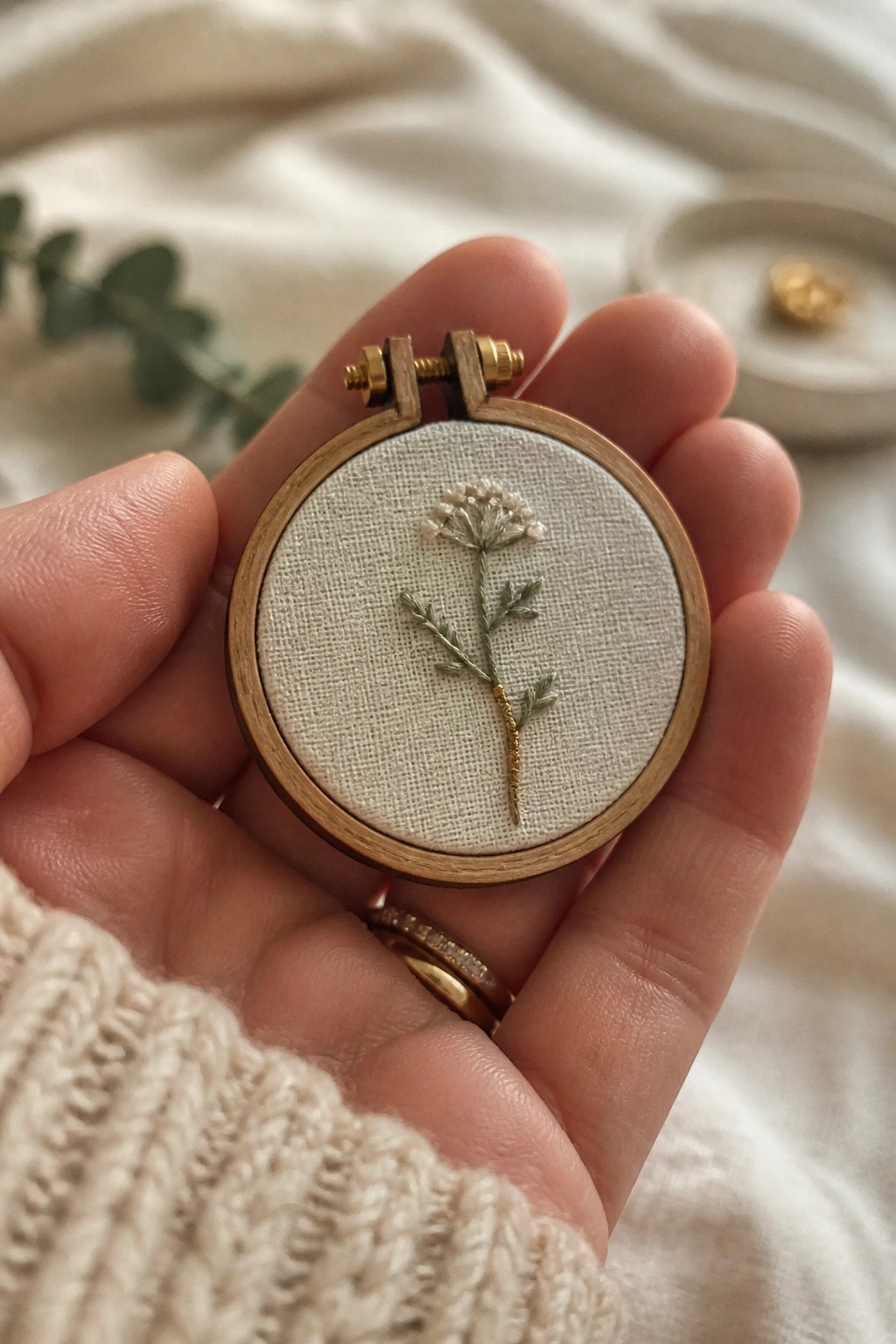 Hand in cream knit sweater holds tiny hoop with green floral embroidery