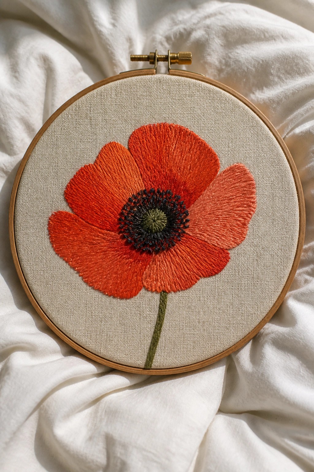 Vibrant red embroidered poppy with black center in wooden hoop on white fabric