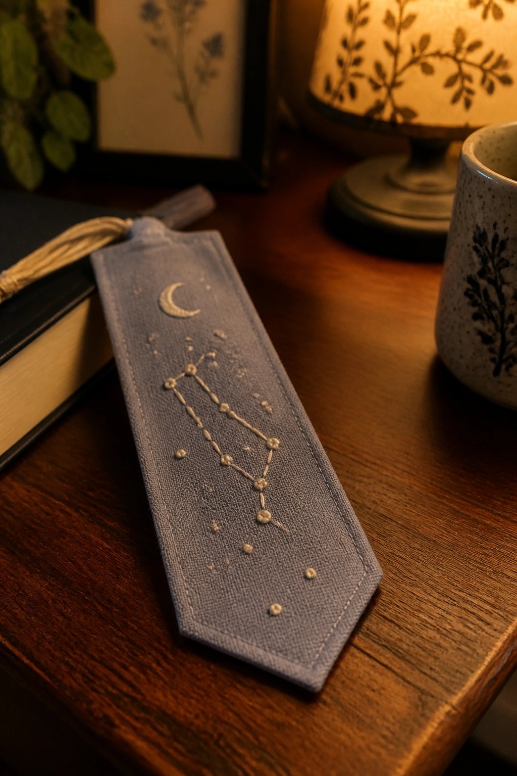 Blue embroidered bookmark with constellation and moon on wooden table beside book
