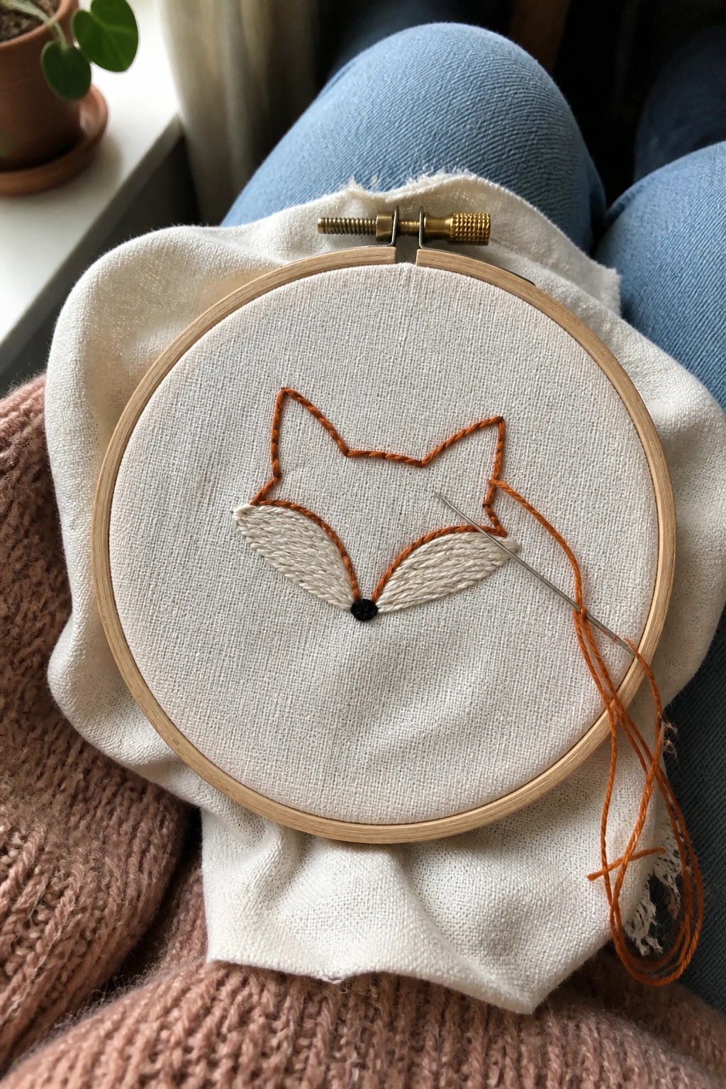 Fox embroidery in orange thread with needle on lap in blue jeans and pink knit