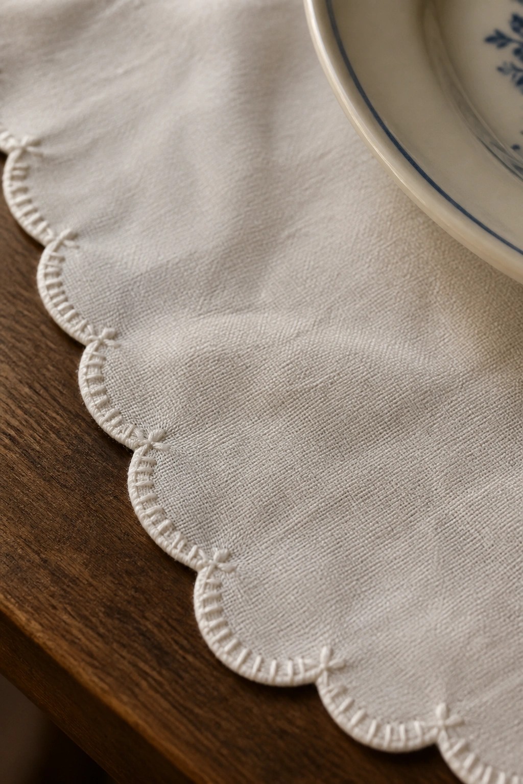White scalloped-edge linen cloth with embroidery on wooden table beside blue-rimmed plate