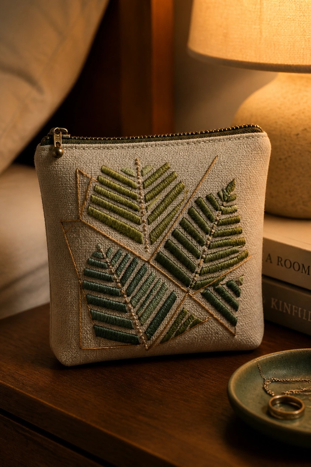 Beige embroidered pouch with green fern leaves on wooden table near lamp and books.