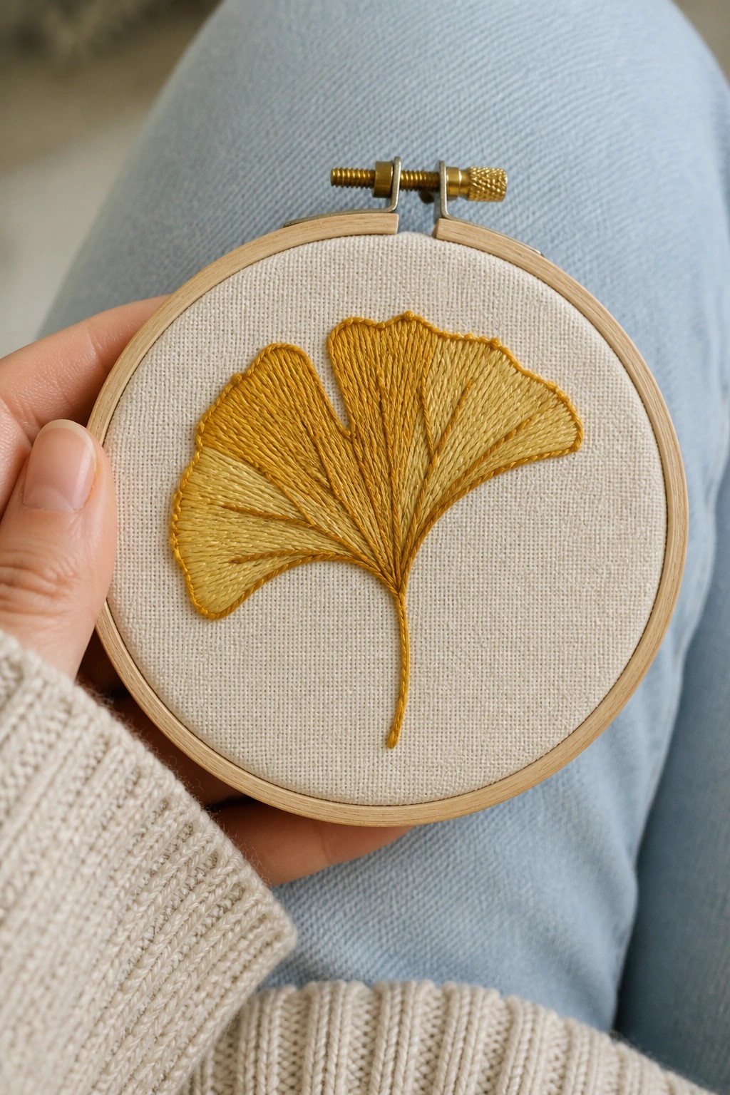 Hand in beige sweater holding hoop with yellow ginkgo leaf embroidery
