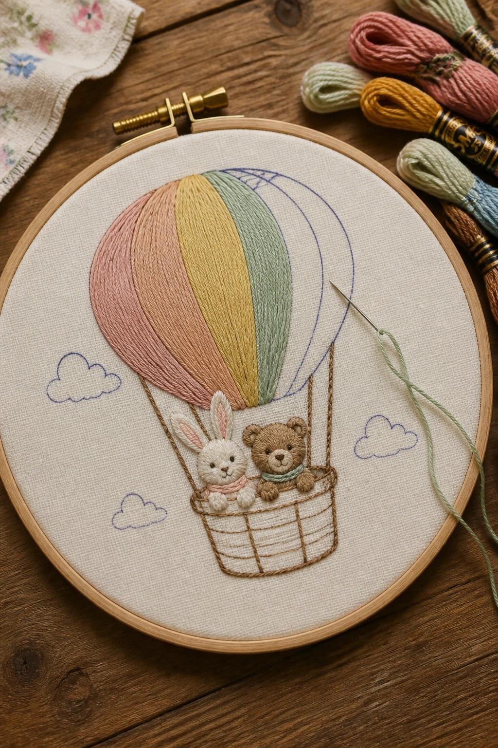 Embroidery hoop with a striped hot air balloon, basket holding a white bunny and brown bear, and blue cloud outlines on white fabric.