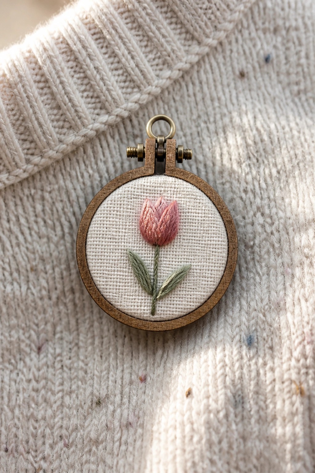 Pink tulip embroidery in wooden hoop on beige knitted sweater.