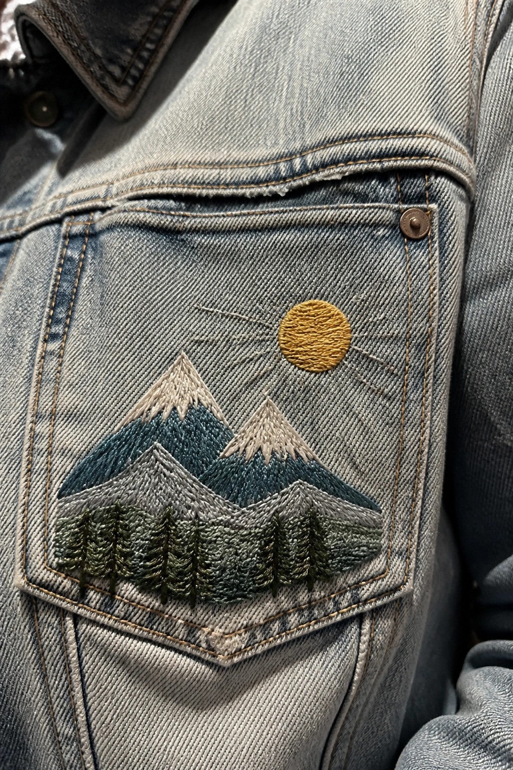 Light blue denim jacket pocket with embroidered mountains, trees, and sun