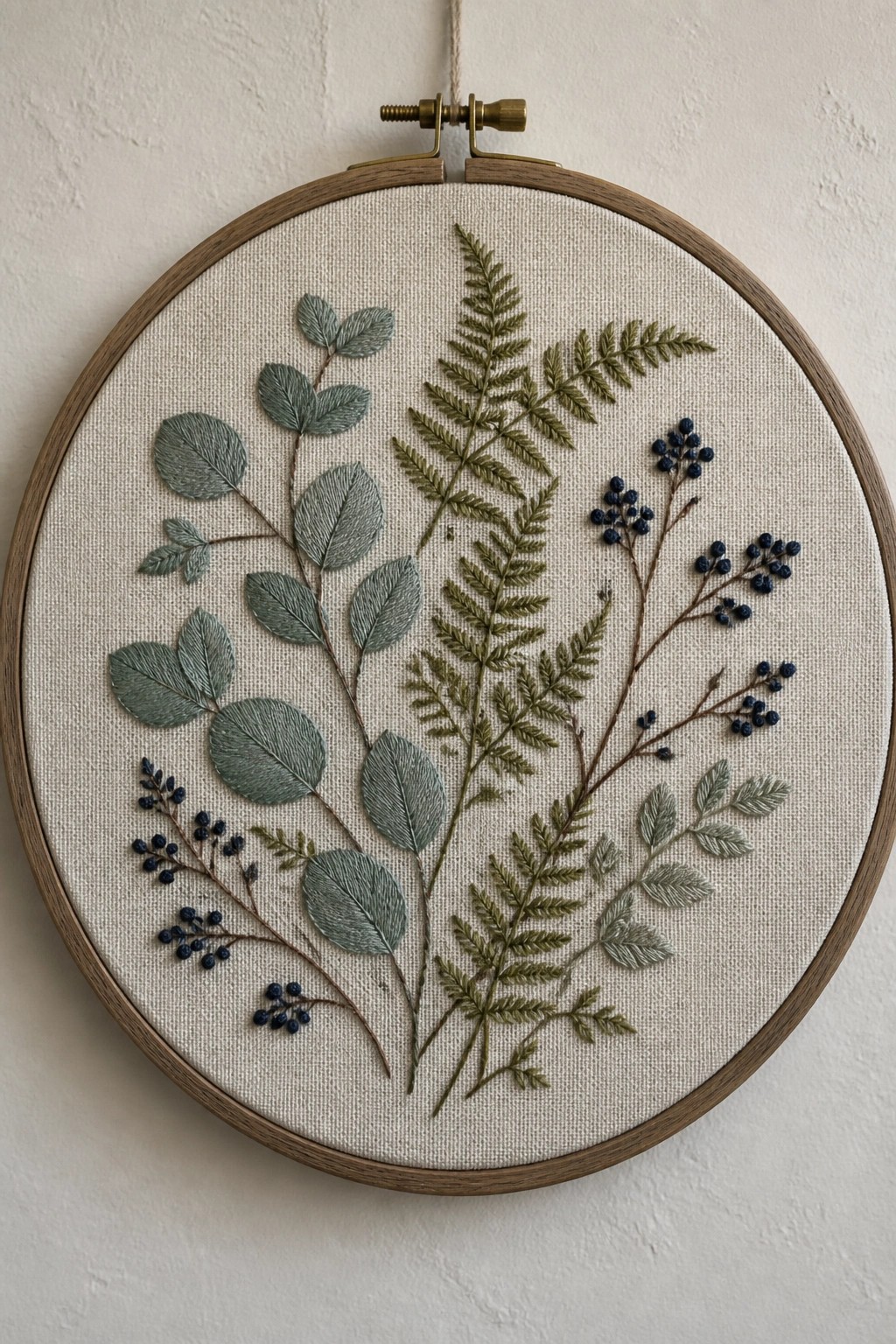 Wooden hoop with embroidered green ferns, eucalyptus leaves, and blue berry clusters.