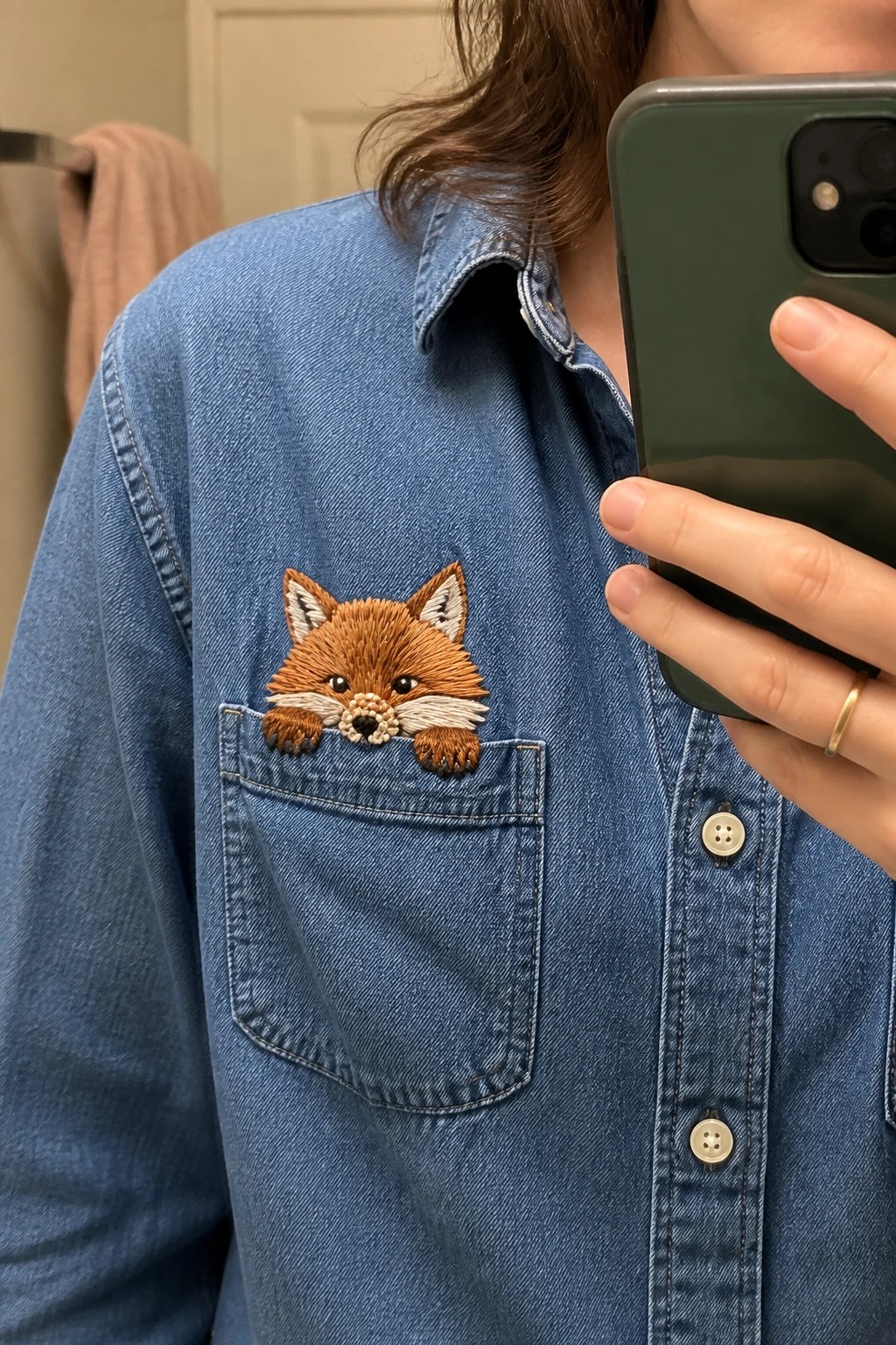 Blue denim shirt with embroidered fox peeking from pocket, hand holding green phone