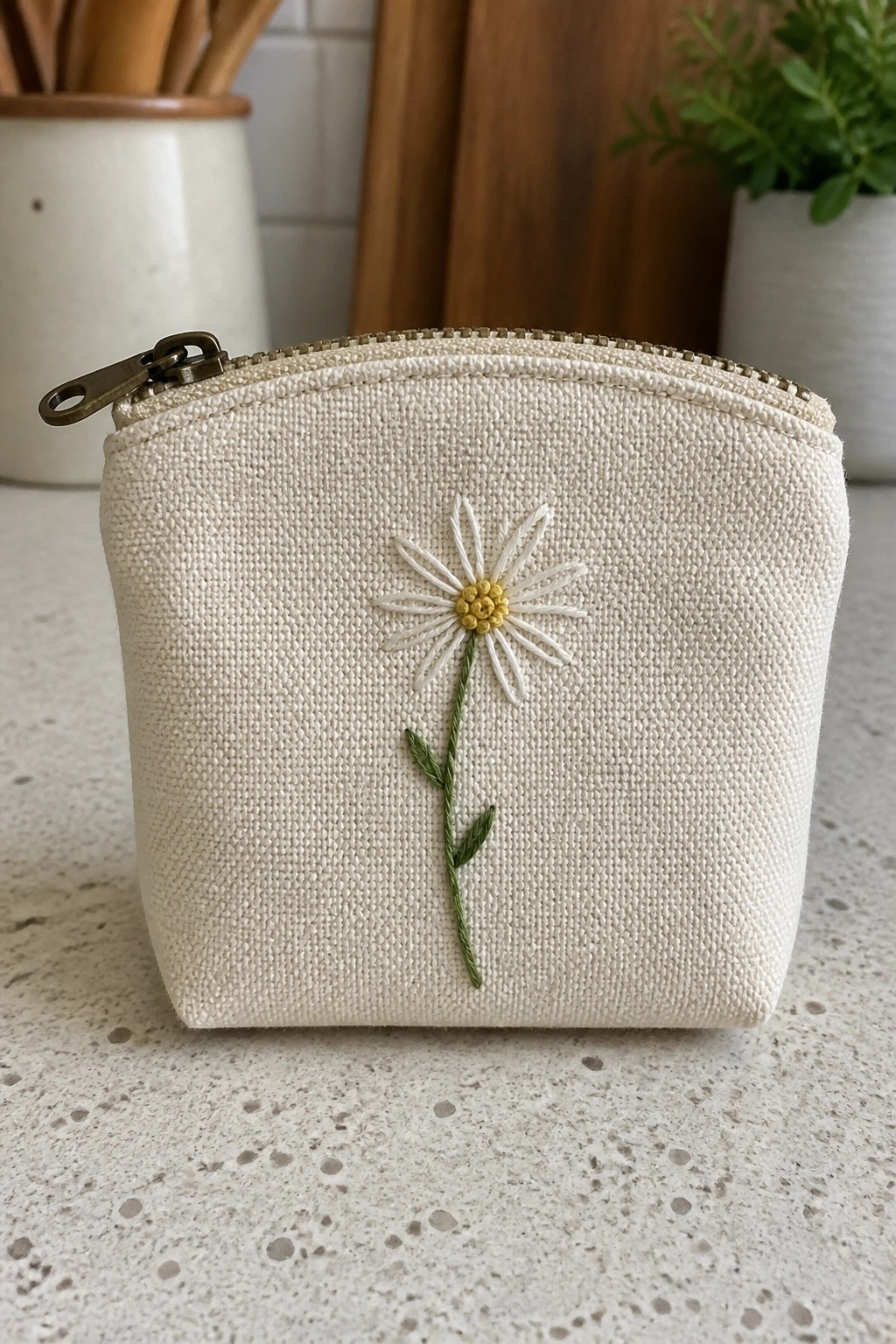 Small off-white fabric pouch with zipper and centered daisy embroidery showing white petals, yellow center, and green stem with leaves.