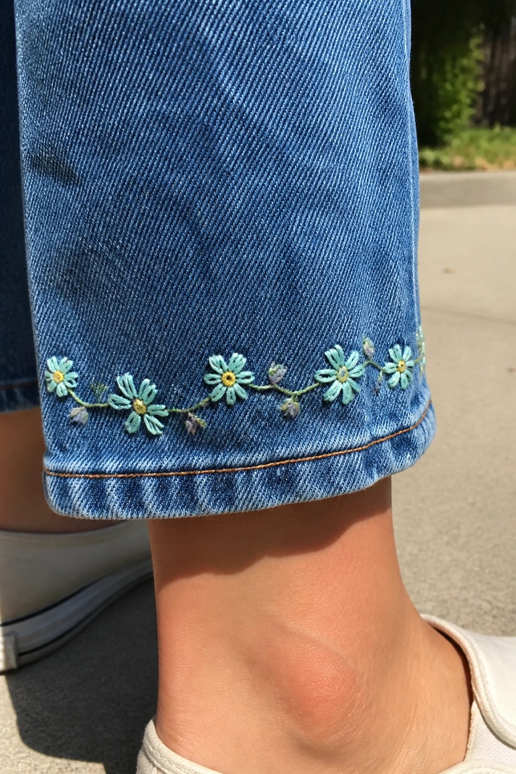 Cuffed blue jeans with light blue daisy embroidery above white sneaker.