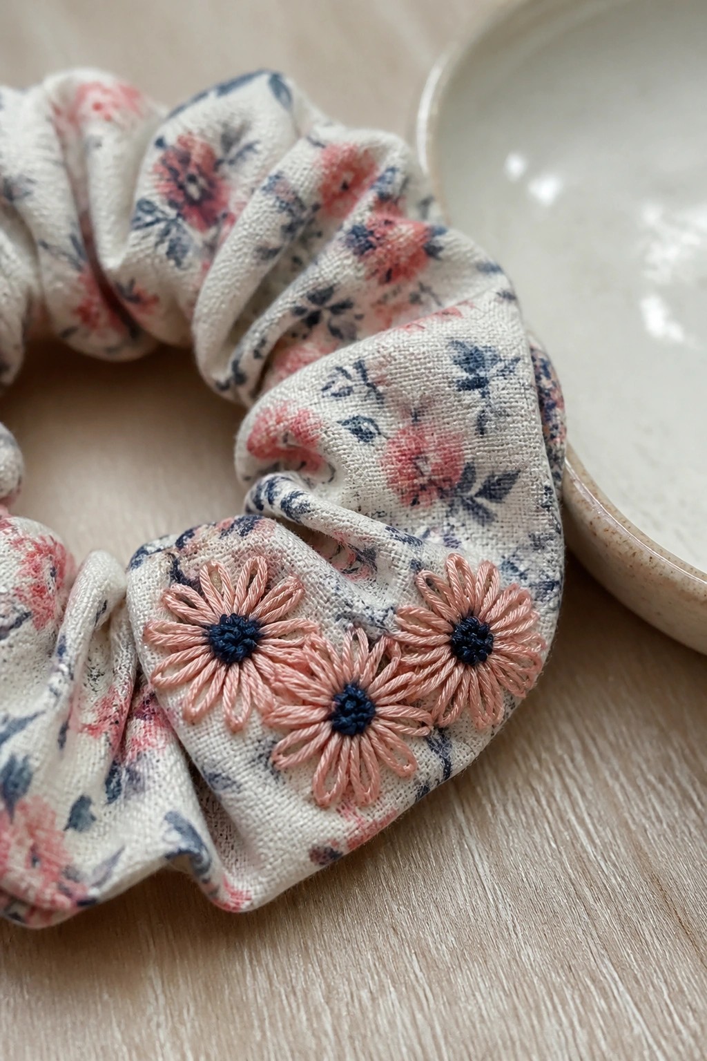 Floral scrunchie with pink embroidered daisies on light wood beside ceramic bowl