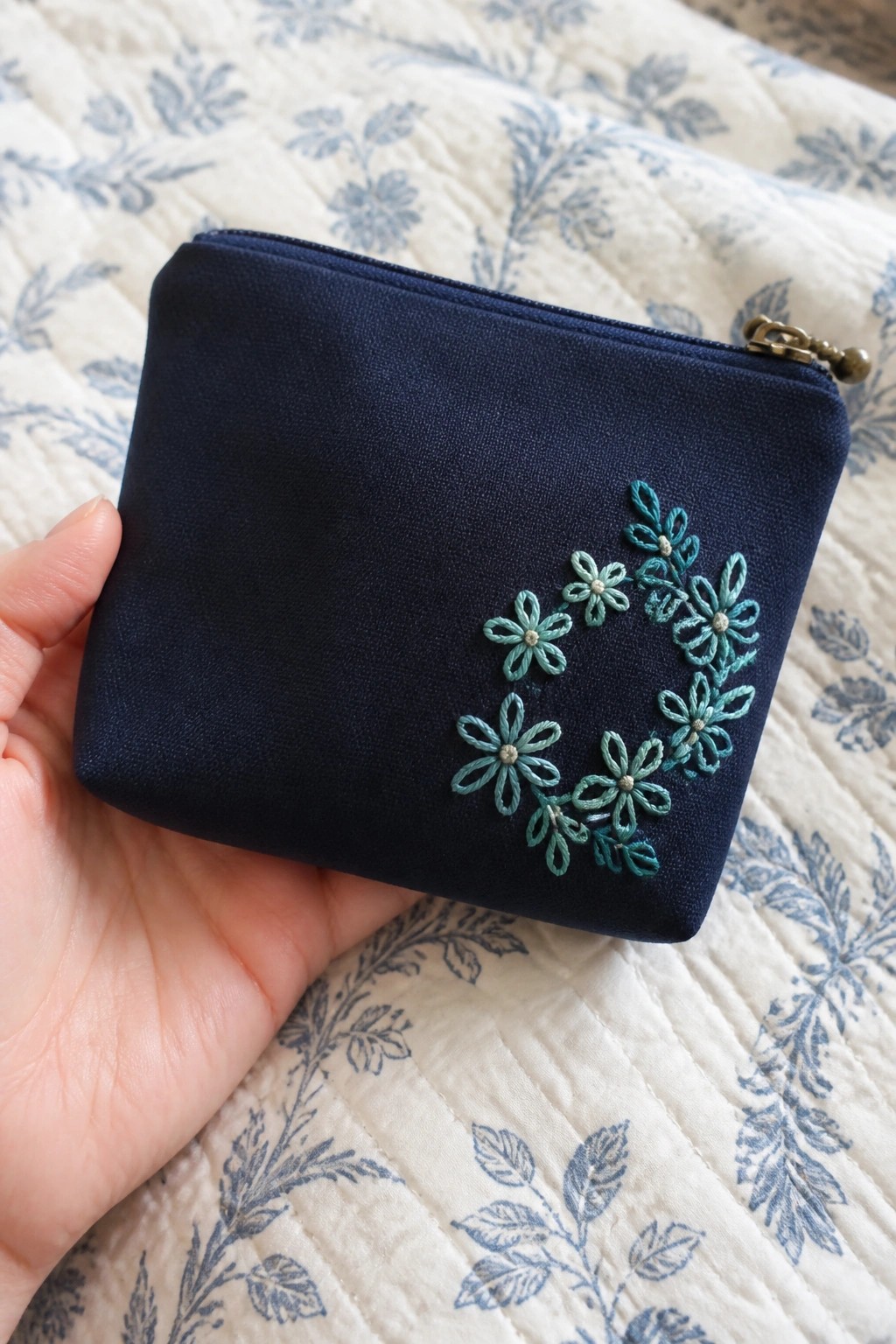 Hand holding navy blue zippered pouch with teal floral embroidery on quilted fabric.