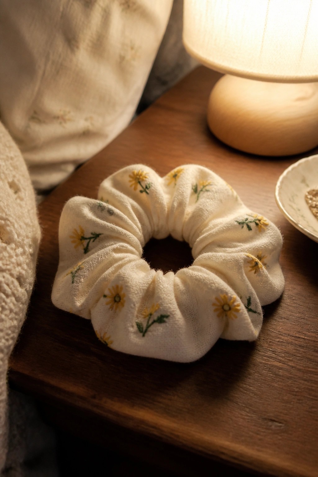 White scrunchie with yellow daisy embroidery on wooden table near lamp