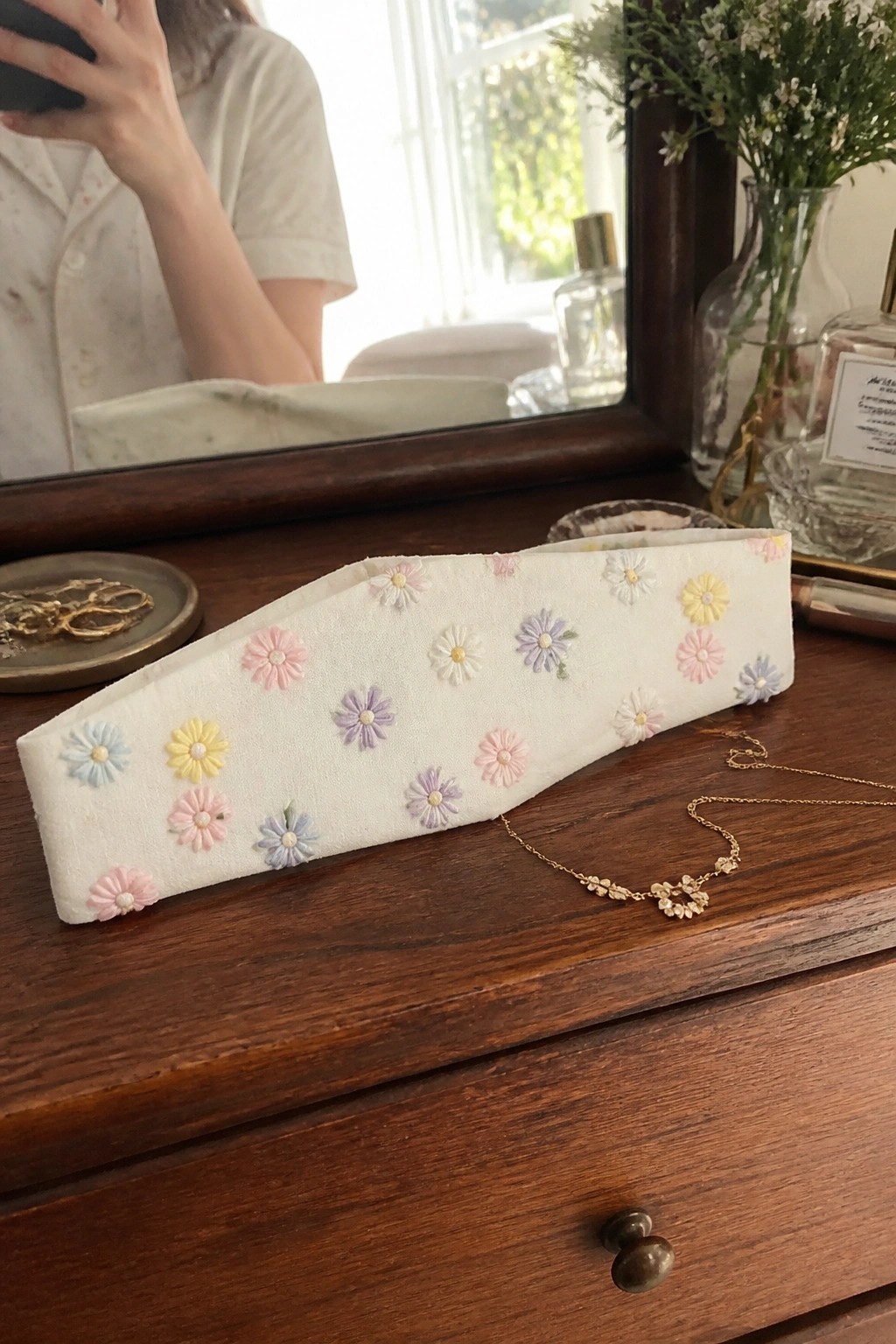 White fabric headband embroidered with small pastel daisy flowers resting on a wooden dresser.