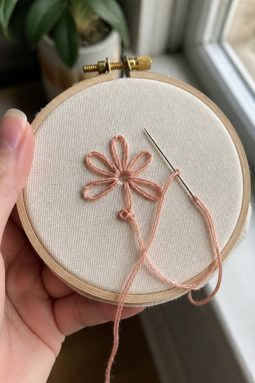 Hand holding wooden hoop with peach embroidered flower, needle, and loose thread on white fabric.