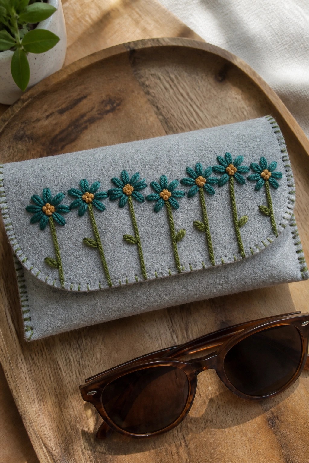 Gray felt pouch with teal embroidered flowers beside tortoiseshell sunglasses on wood.