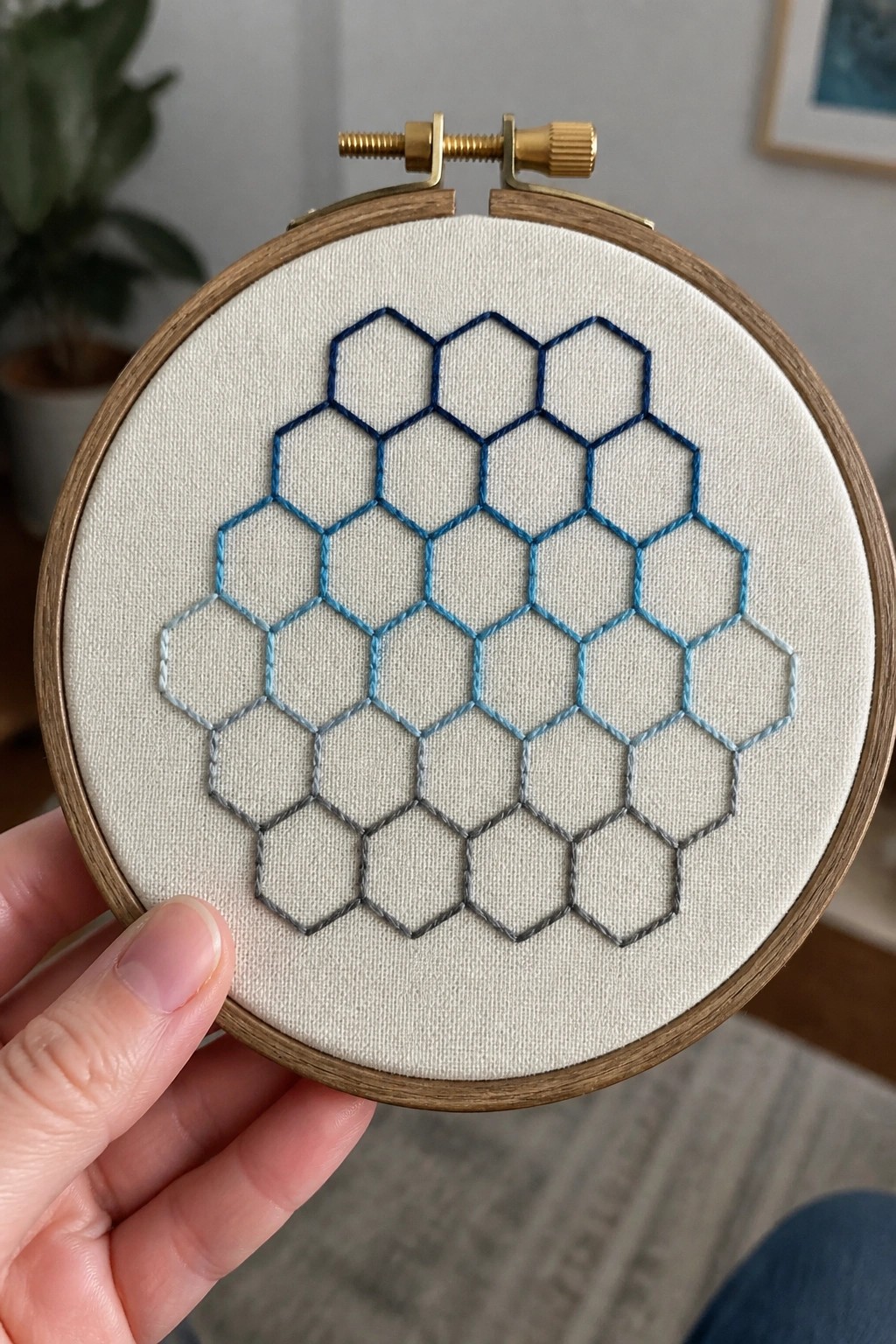 Hand holding wooden hoop with blue-to-gray hexagonal embroidery on white fabric.