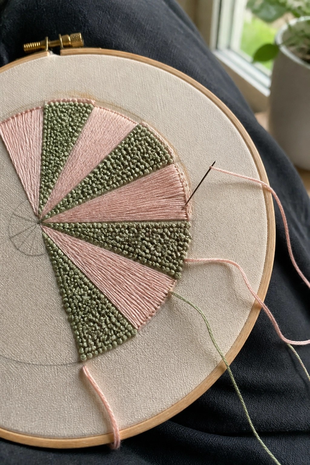 Embroidery hoop with pink and green pie-slice stitches, needle, and loose threads on fabric.