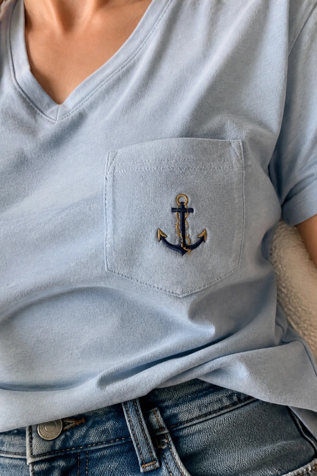 Light blue V-neck t-shirt with navy and gold embroidered anchor on chest pocket over jeans