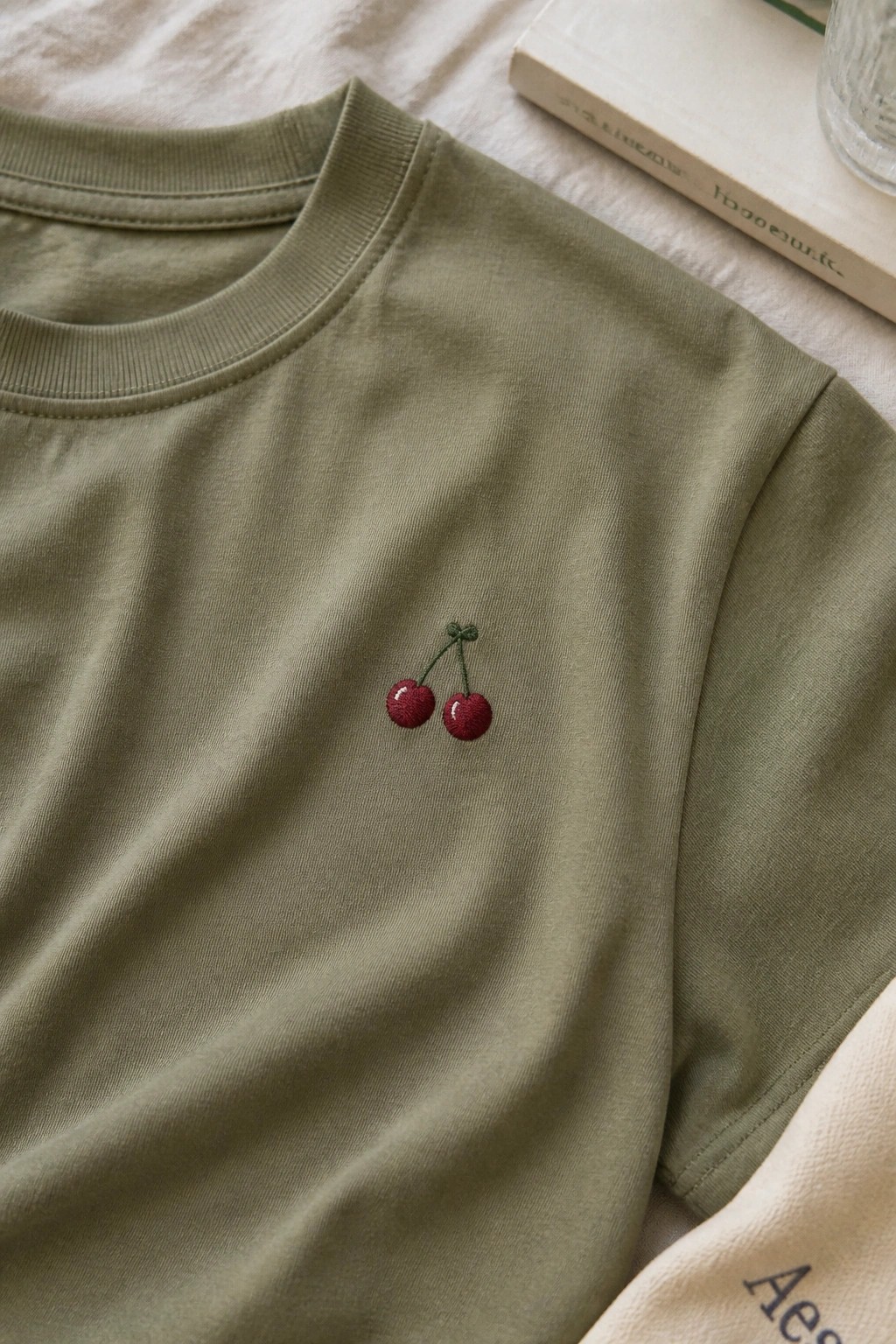 Sage green t-shirt with cherry embroidery beside a book on white fabric.