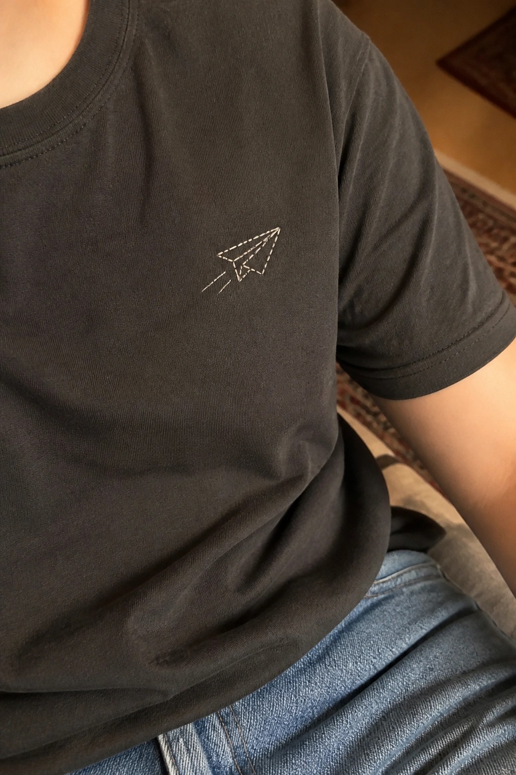 Black t-shirt with small white paper airplane embroidery on the left chest.