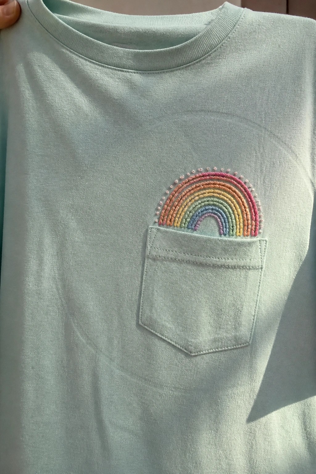 Mint green t-shirt with embroidered rainbow above front pocket, held by hand