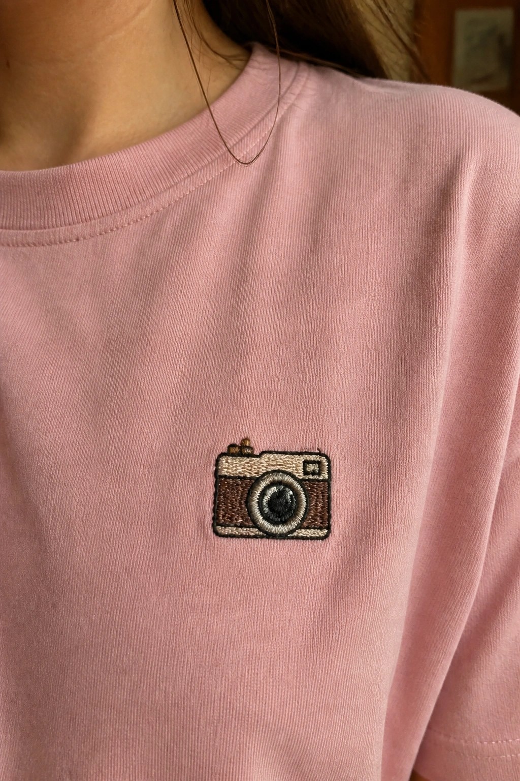 Light pink t-shirt with embroidered brown camera patch on the chest.