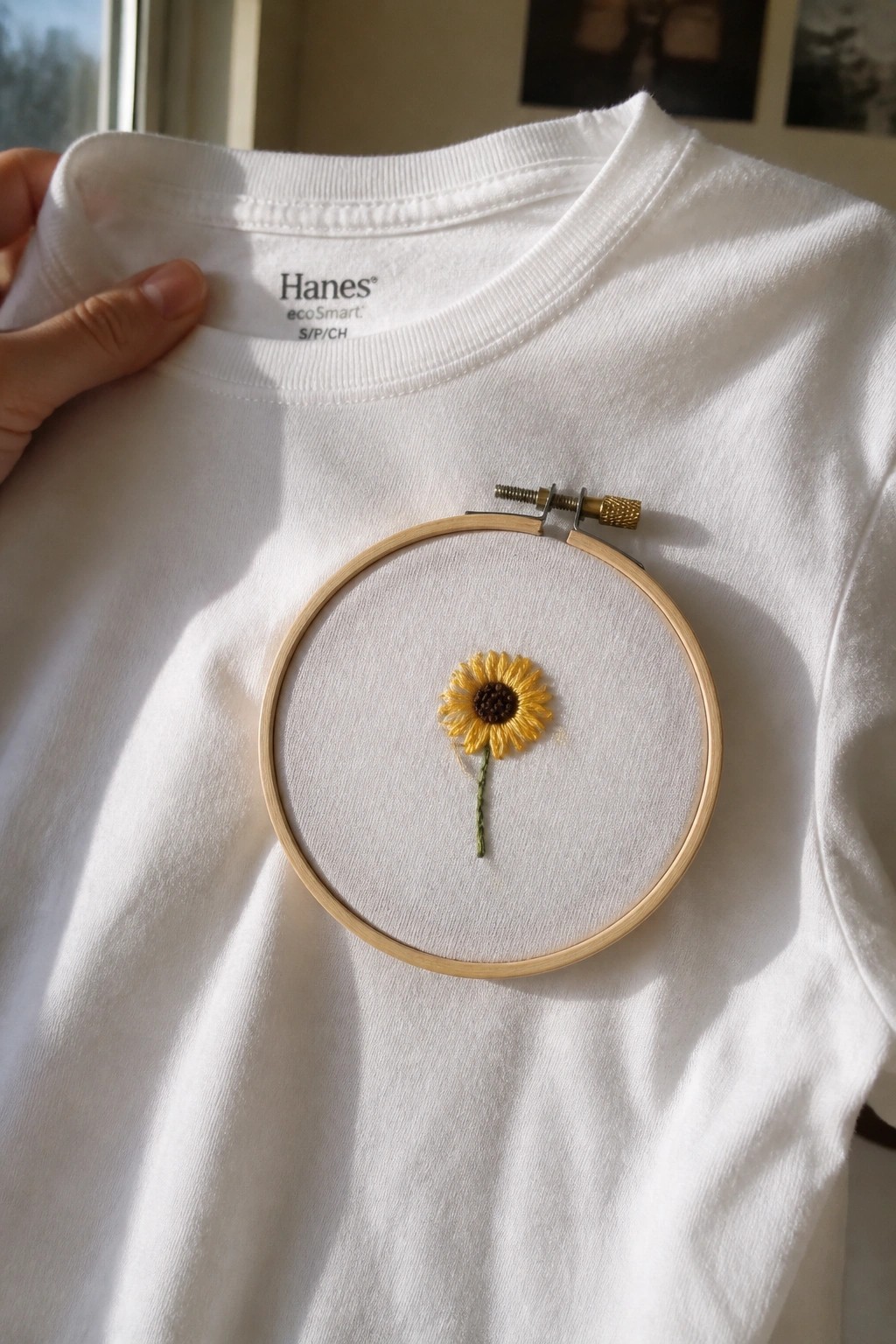 White t-shirt with yellow sunflower embroidery in wooden hoop held by hand.