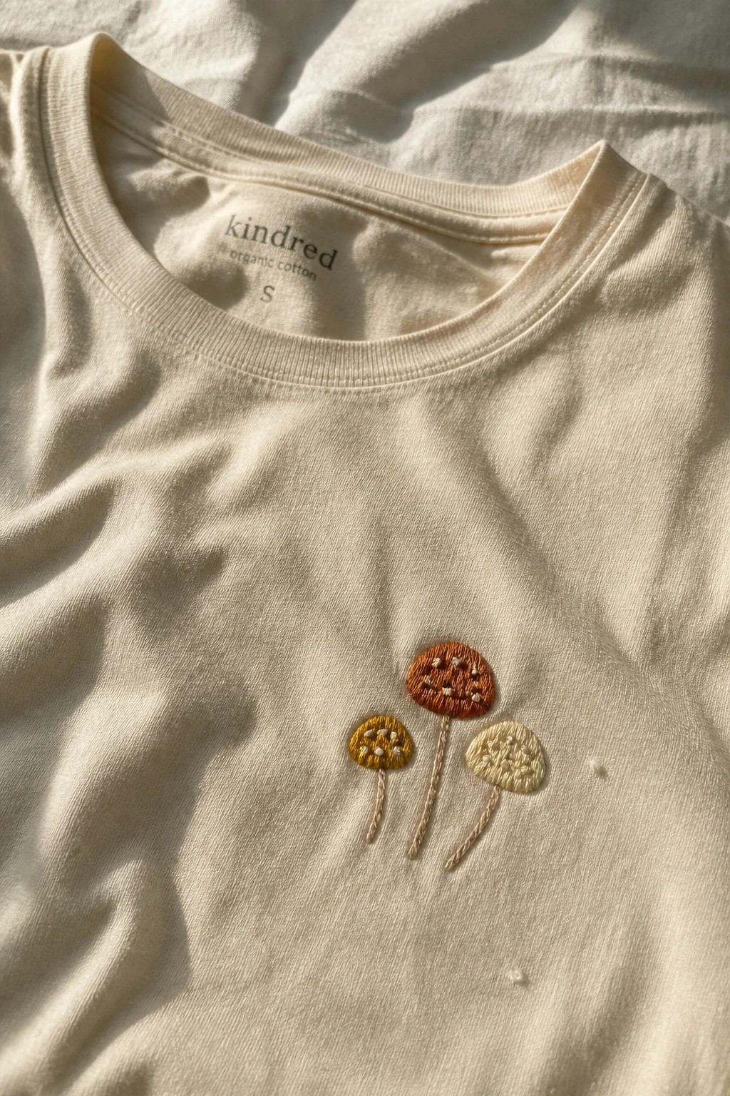 Close-up of a cream t-shirt with three small embroidered mushrooms on the front chest.