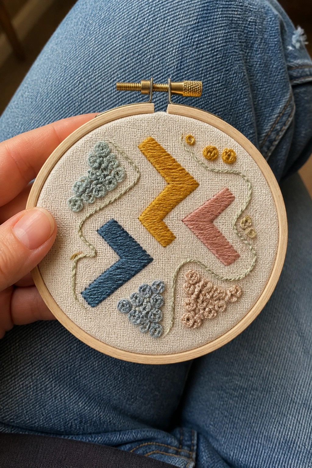 Embroidery hoop with blue, yellow, and pink chevron shapes plus small rosette clusters in blue, yellow, and beige connected by green threads on beige fabric.