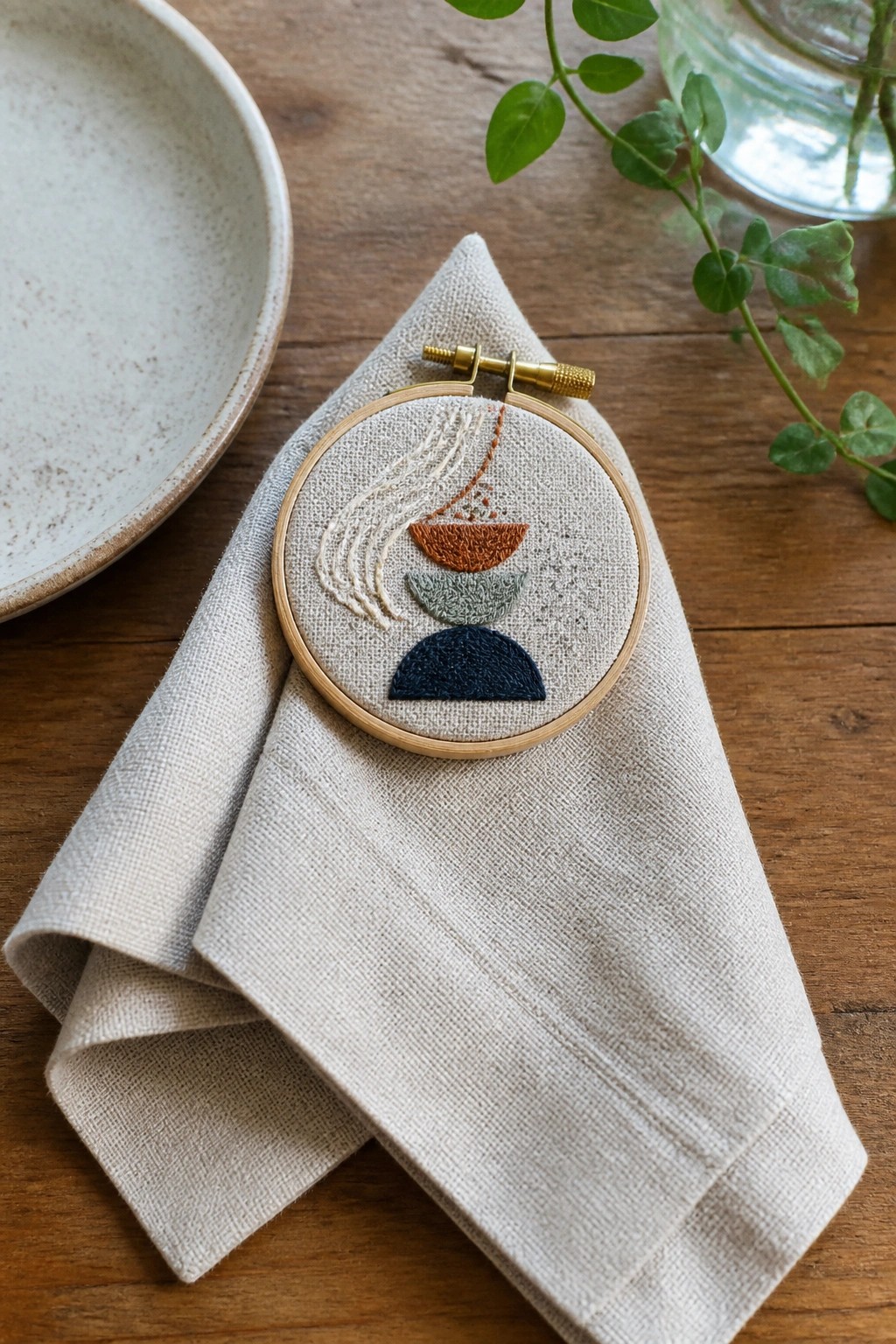 Abstract embroidery hoop on beige linen beside white plate and green plant on wood