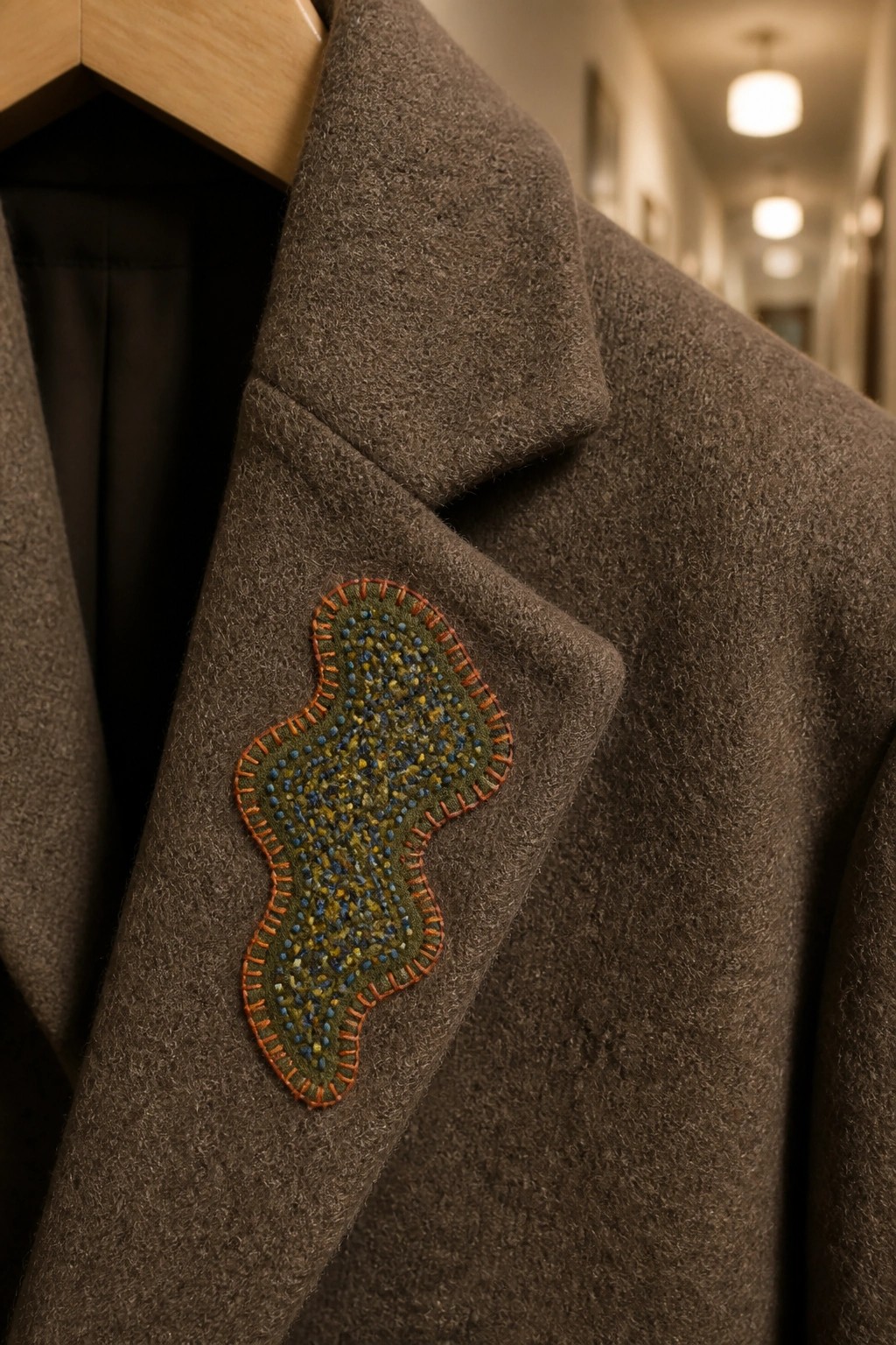 Brown wool coat with green embroidered patch edged in orange on wooden hanger