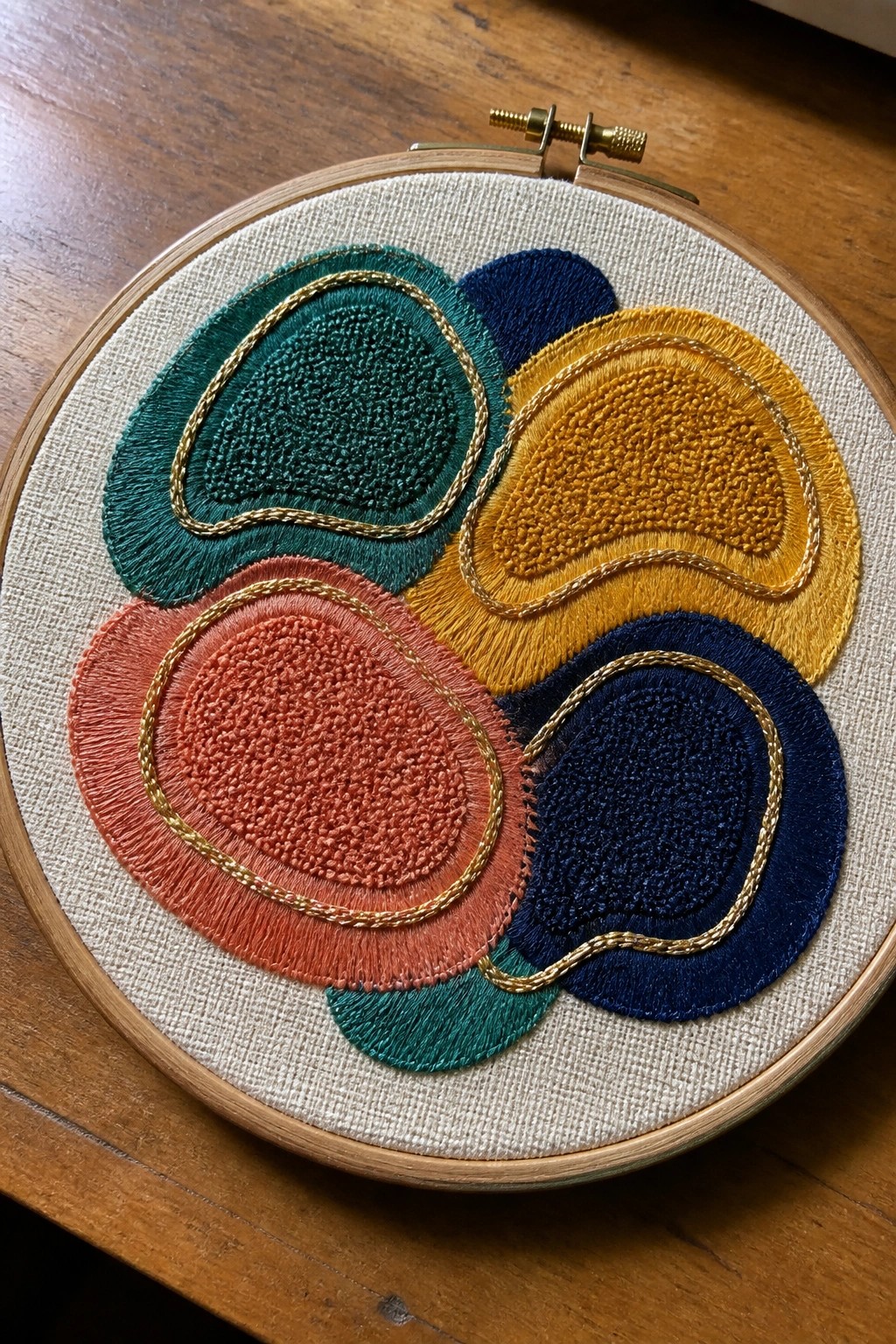 Colorful abstract embroidery in wooden hoop with gold outlines on beige fabric.