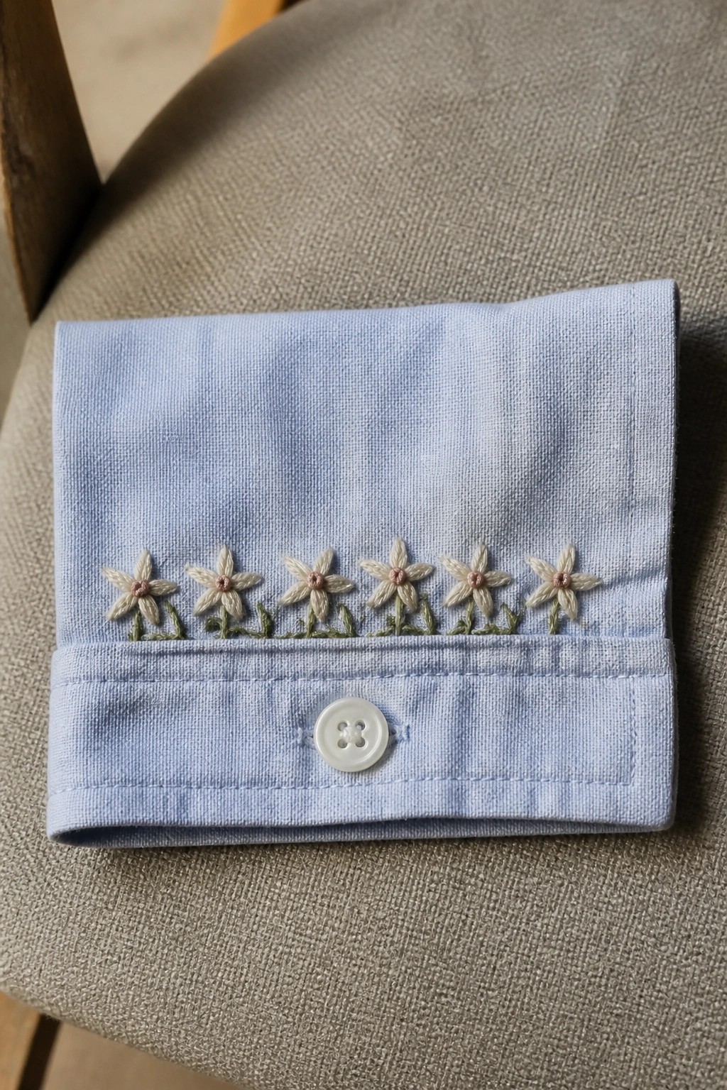 Light blue shirt cuff with white daisy embroidery, pink centers, and button on gray fabric.