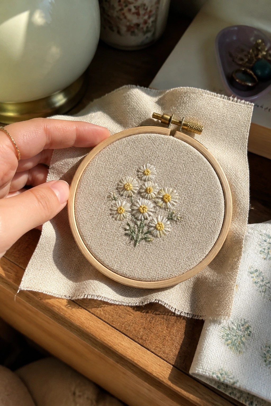 A small wooden embroidery hoop holds a cluster of white daisies with yellow centers and green stems on beige fabric.