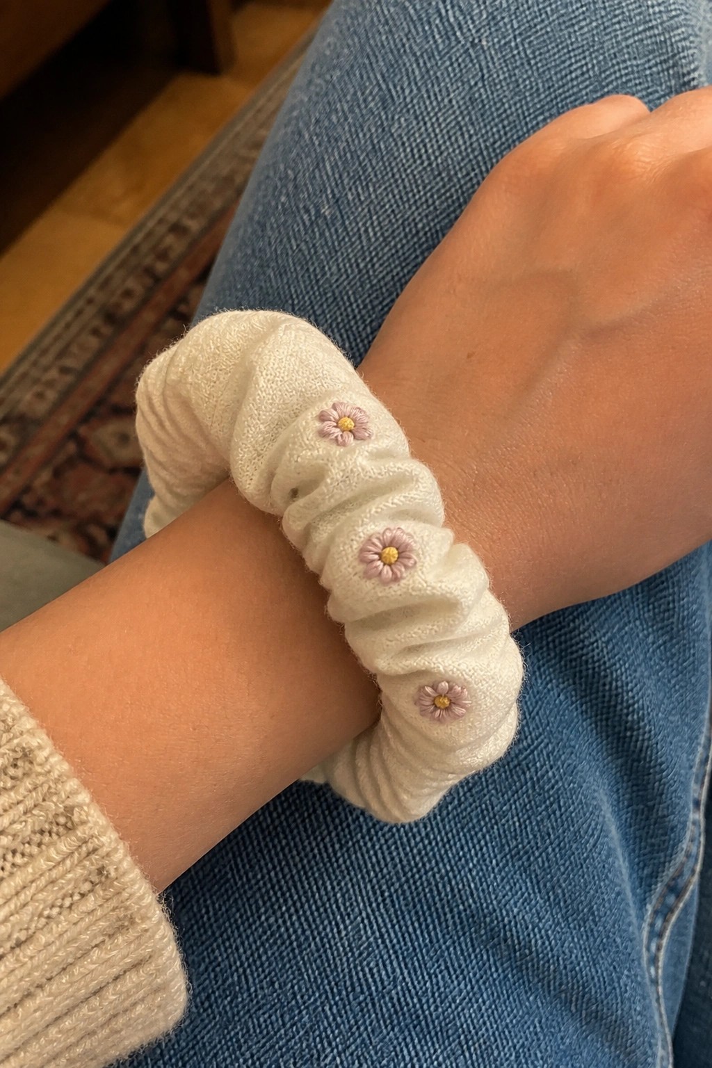 White scrunchie with pink daisy embroidery on wrist over blue denim jeans
