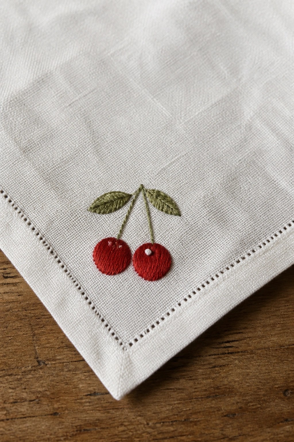 White napkin corner with two red embroidered cherries, green leaves, and stems on wood.
