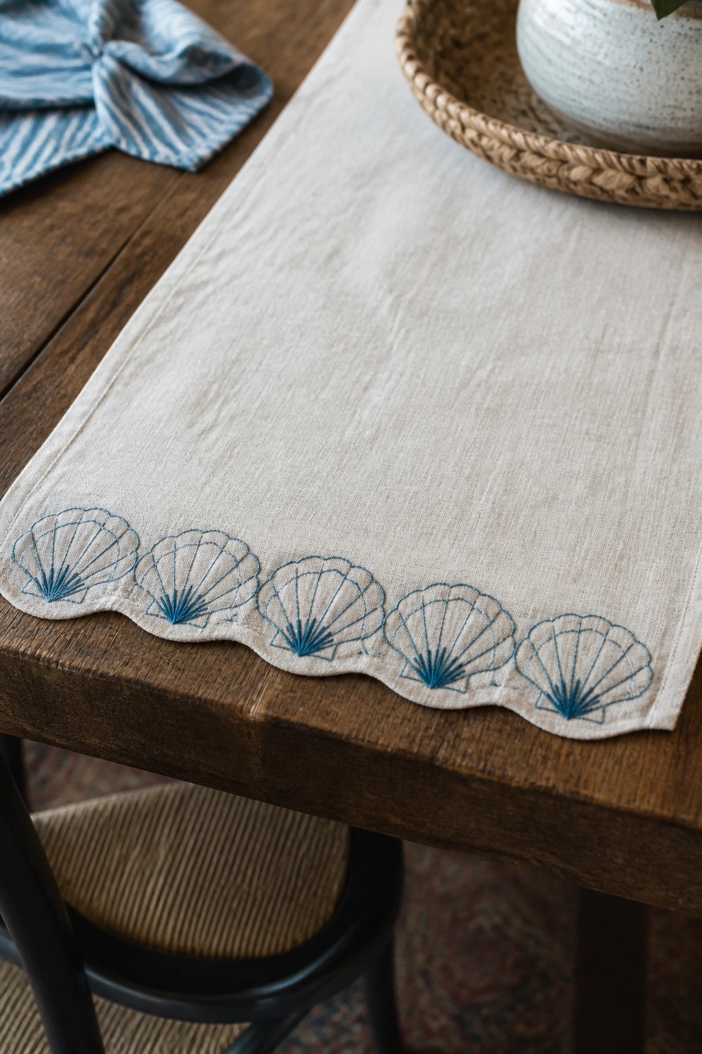 A close-up of a linen table runner featuring five blue embroidered seashells along its scalloped bottom edge on a wooden table.