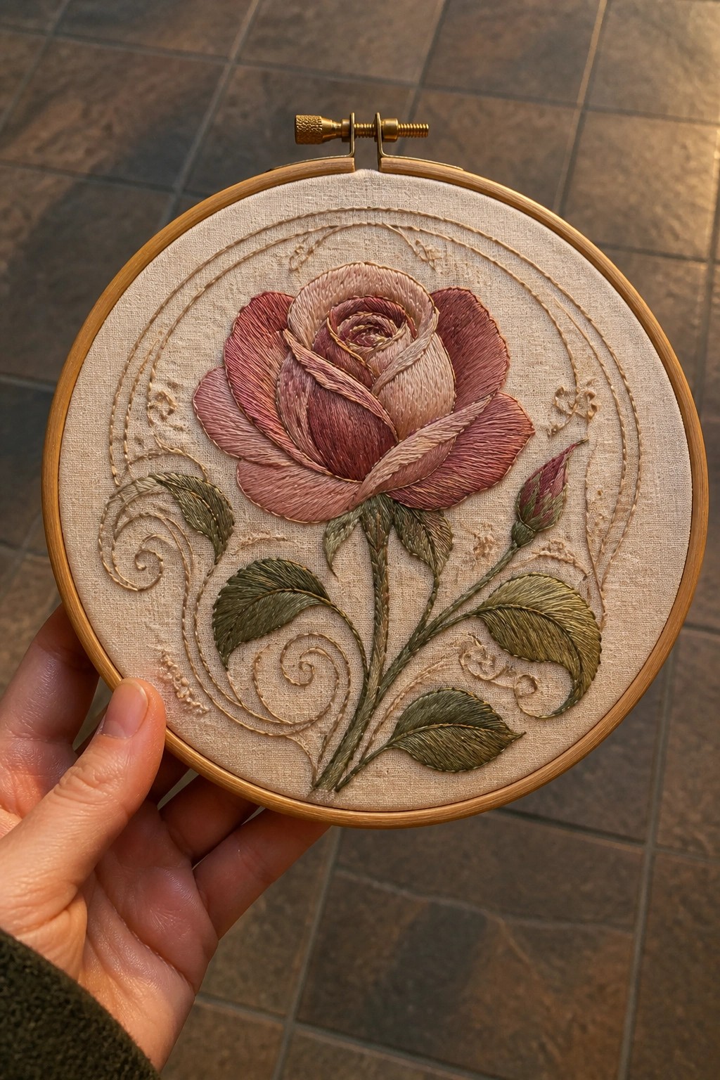 Hand holding wooden hoop with embroidered pink rose, green leaves on beige fabric